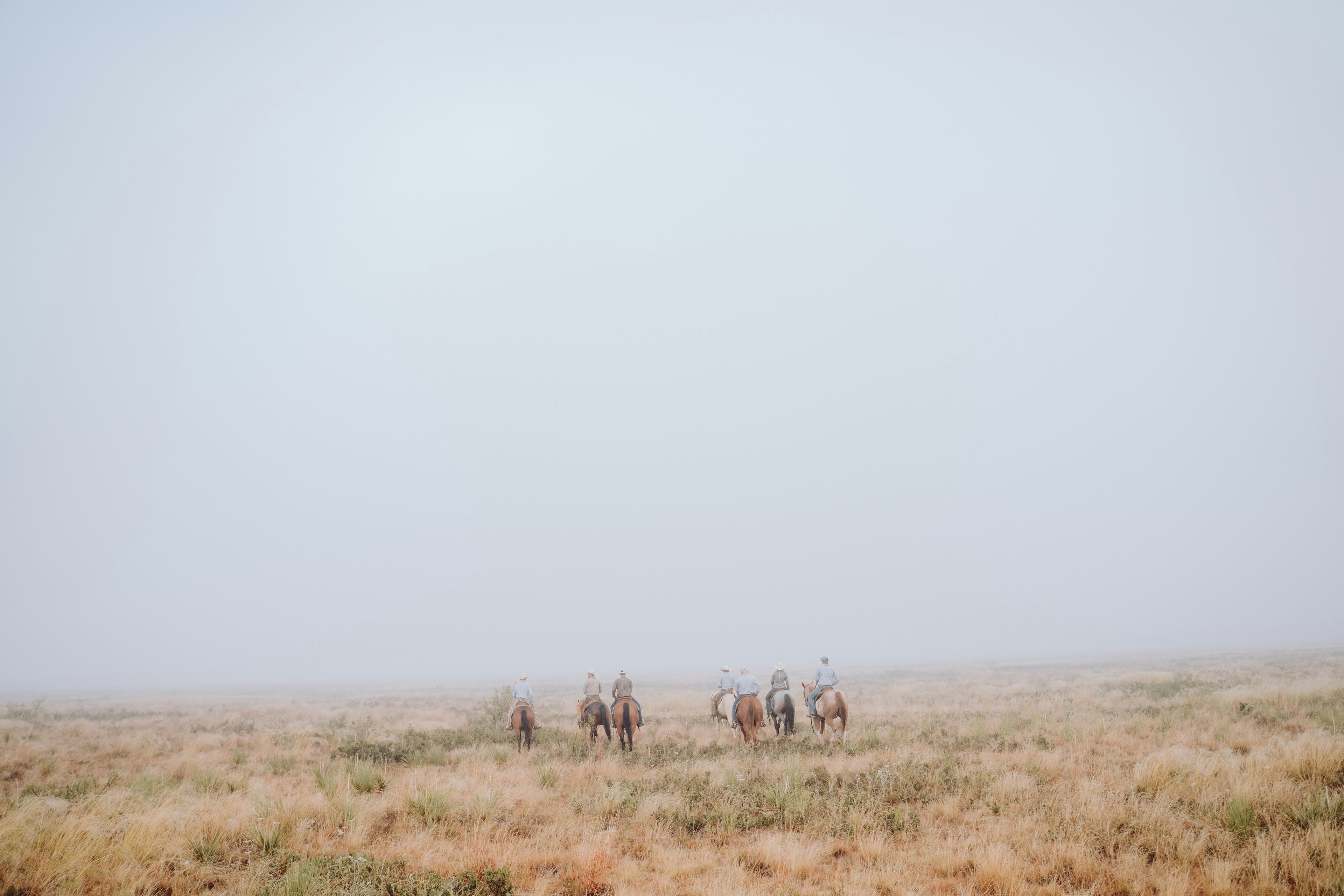 Multiple people on horse back riding away on a foggy prairie
